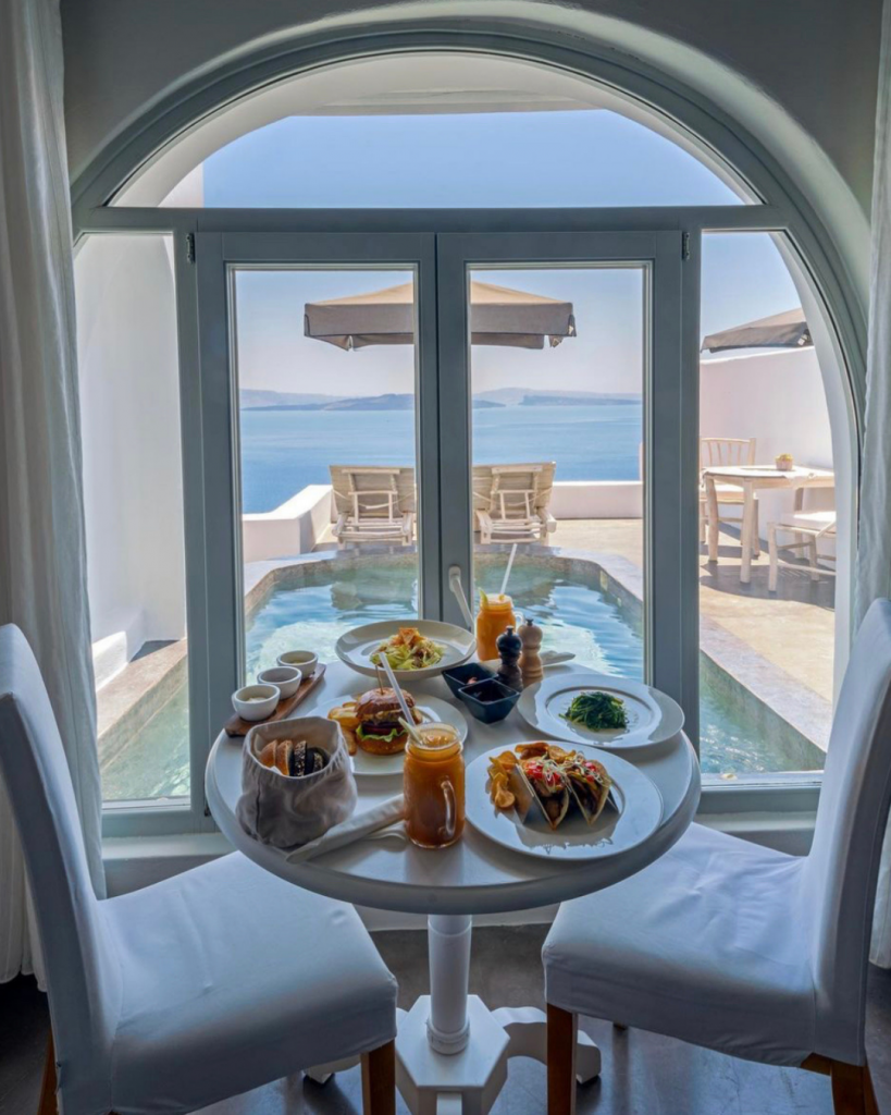A table with 2 chairs. Breakfast is served on the table and in the background is a balcony with a pool. Pythia Suite in Andronis Luxury Suites in Santorini